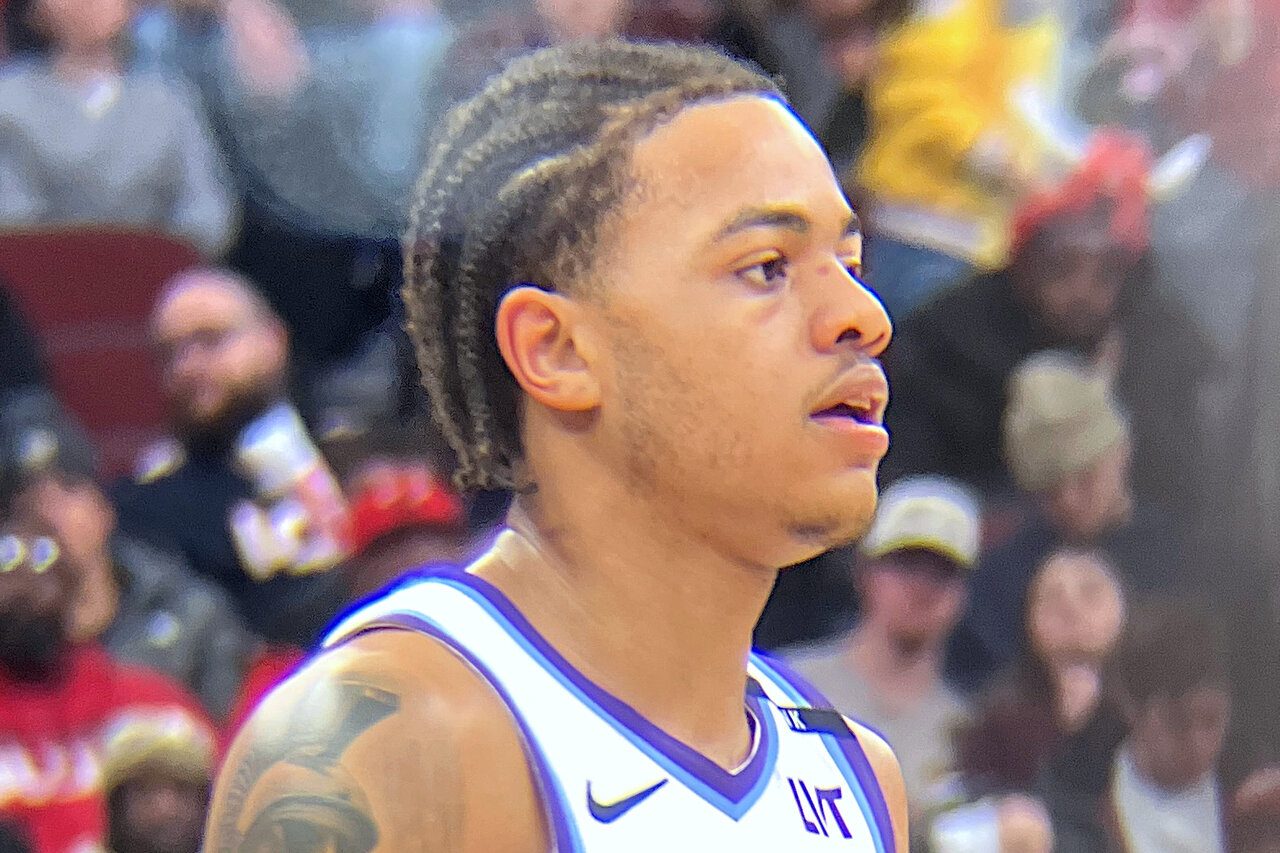 Keyonte George in a white Utah Jazz jersey with purple mountain graphics, braided hair and a tattooed left arm, looking to his right with fans blurred behind him