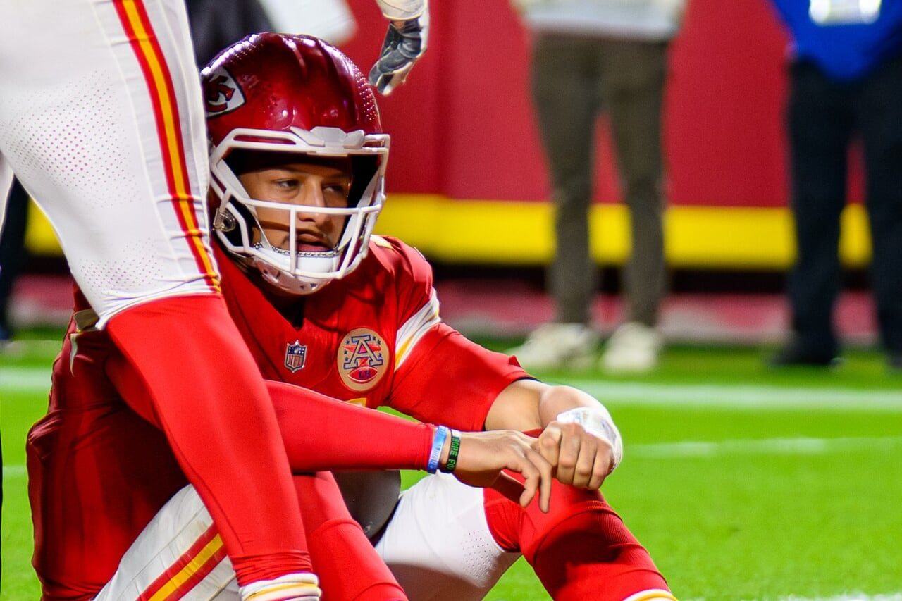Patrick Mahomes in a red Kansas City Chiefs helmet and uniform sits on the field after a play as a teammate stands over him