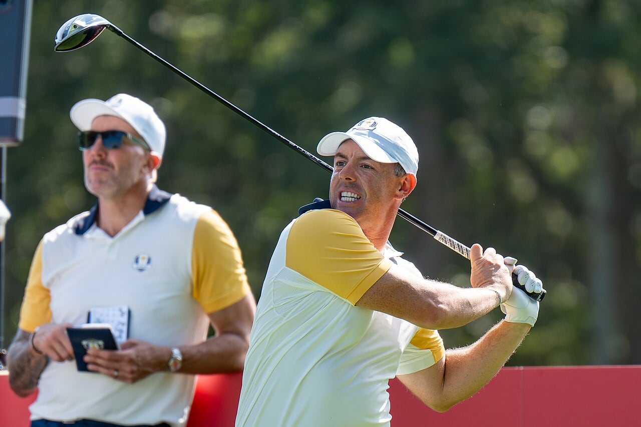 Rory McIlroy in a white cap and a white and yellow Europe team shirt watches his tee shot at the 2025 Ryder Cup, with a man in matching team clothing behind him