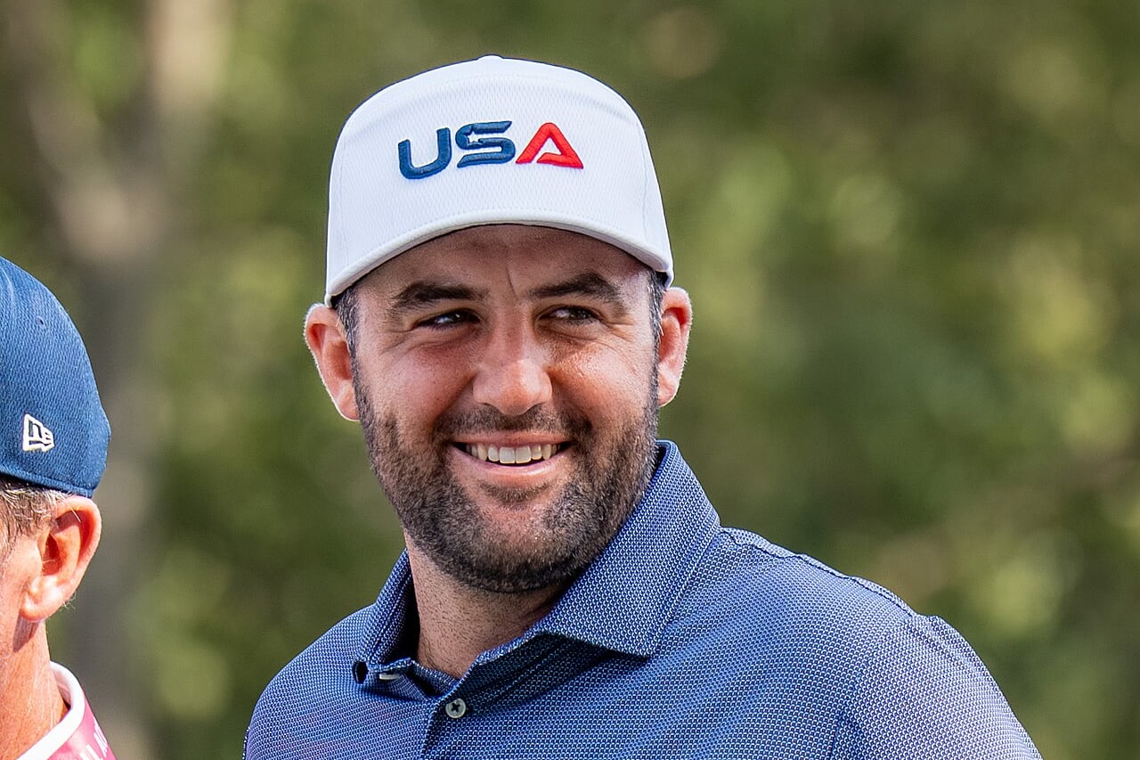 Scottie Scheffler smiling in a white USA cap and a navy Ryder Cup polo shirt, with trees blurred behind him