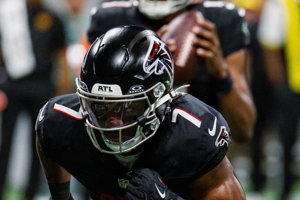 Bijan Robinson in a black Atlanta Falcons helmet and number 7 jersey crouches in a three-point stance, with a teammate holding a football behind him