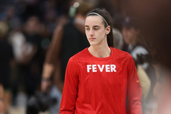 Caitlin Clark in a red Indiana Fever warm-up shirt looks down as she walks onto the court before a game