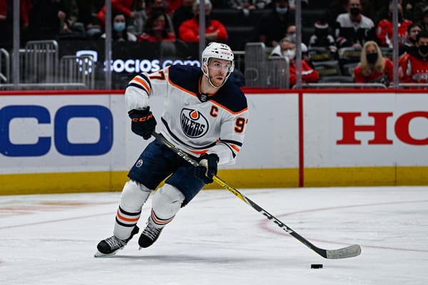 Connor McDavid in a white Edmonton Oilers road jersey with the captain's C skates with the puck in front of the boards