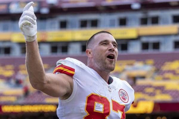 Travis Kelce in a white Kansas City Chiefs jersey, number 87, points skyward and shouts toward the stands