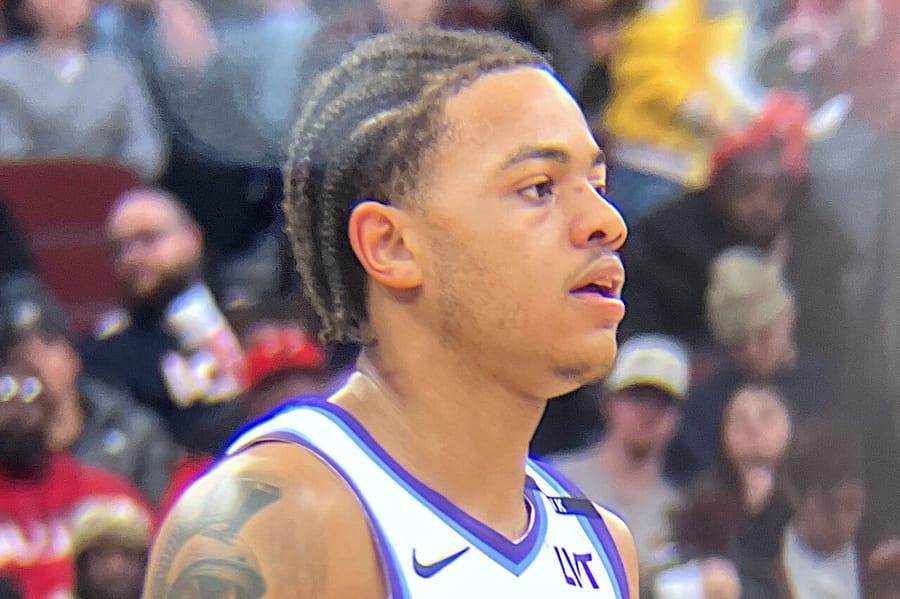 Keyonte George in a white Utah Jazz jersey with purple mountain graphics, braided hair and a tattooed left arm, looking to his right with fans blurred behind him