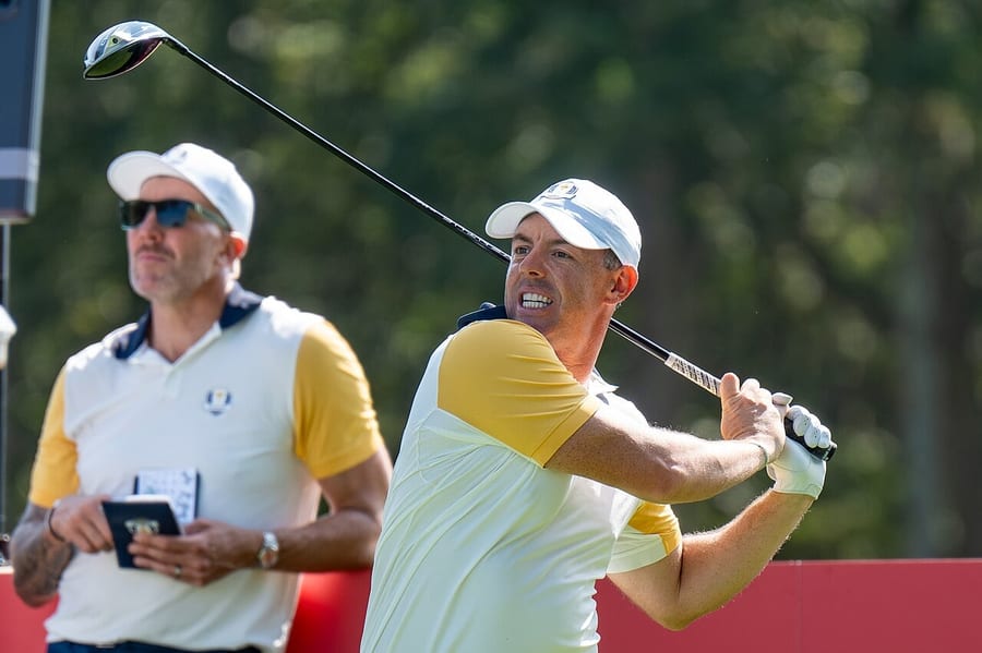 Rory McIlroy in a white cap and a white and yellow Europe team shirt watches his tee shot at the 2025 Ryder Cup, with a man in matching team clothing behind him