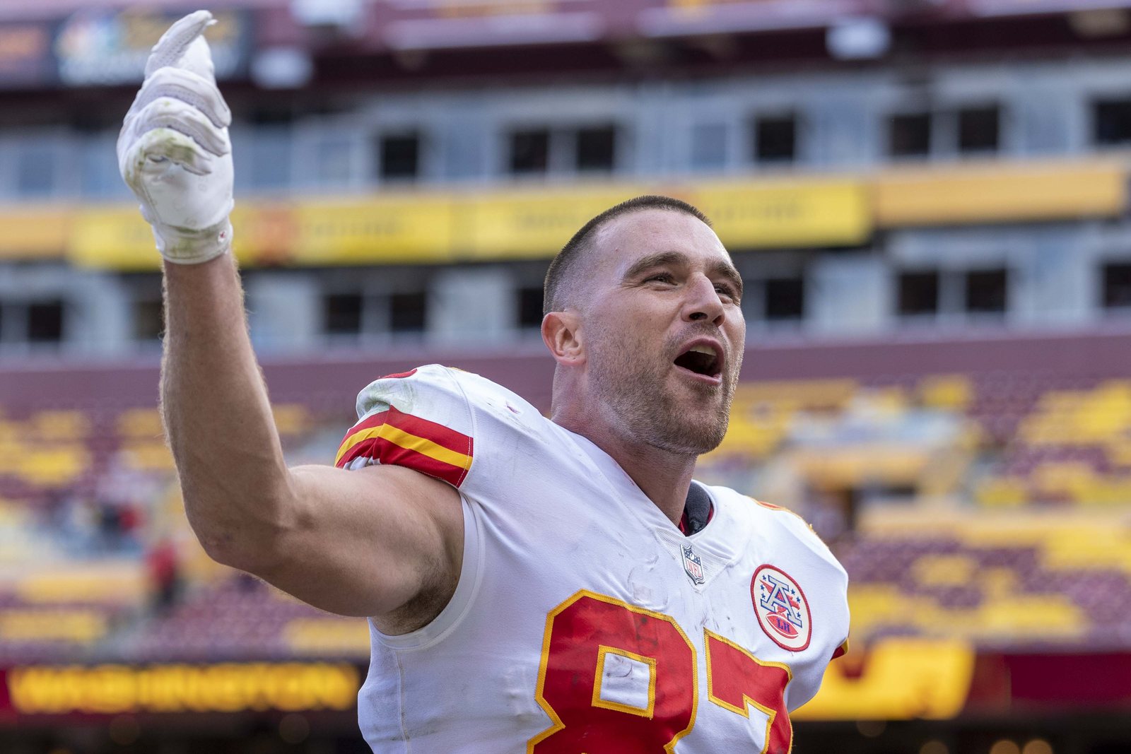 Travis Kelce in a white Kansas City Chiefs jersey, number 87, points skyward and shouts toward the stands